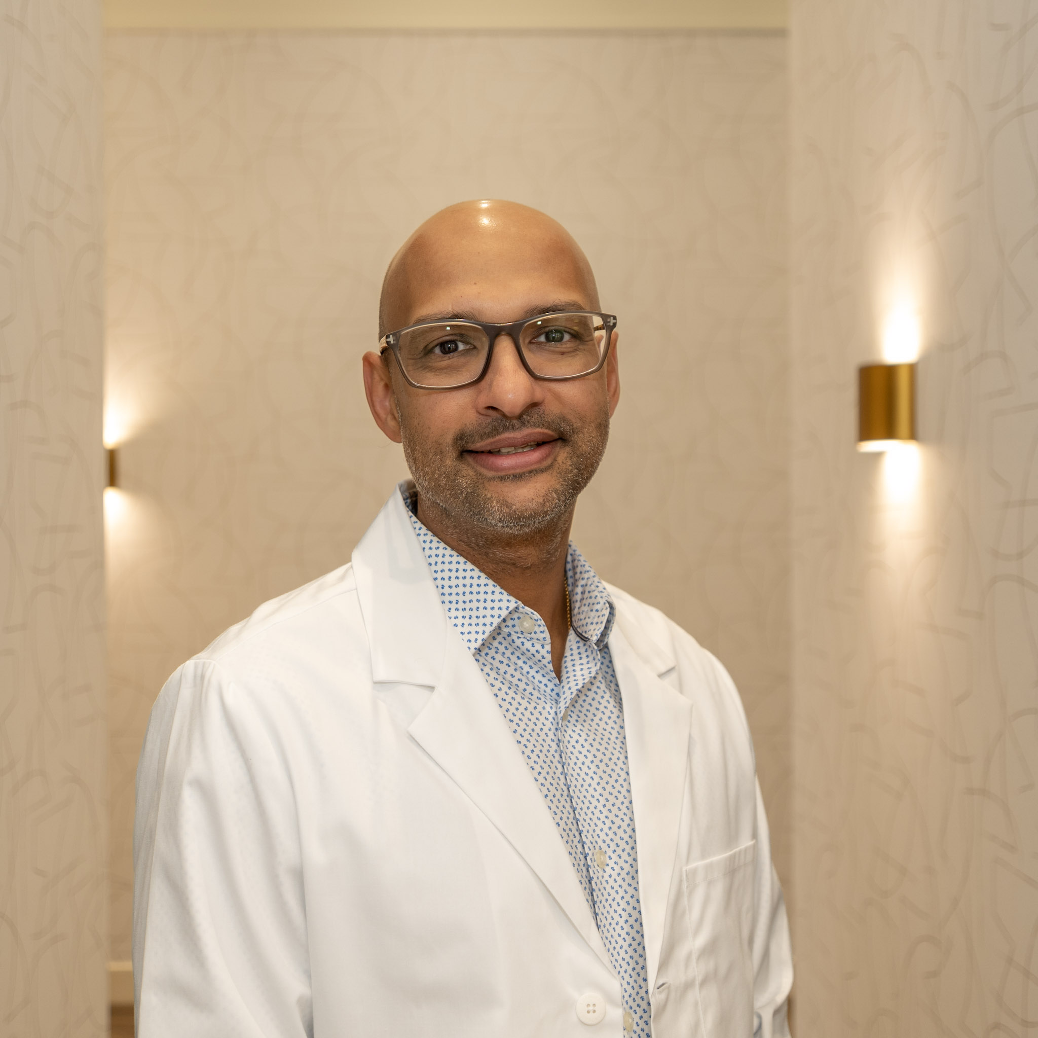 Dr. Ashish Oommen, a general and restorative dentist at Spray Lake Dental in Cochrane, Alberta, smiling in a white lab coat and glasses.