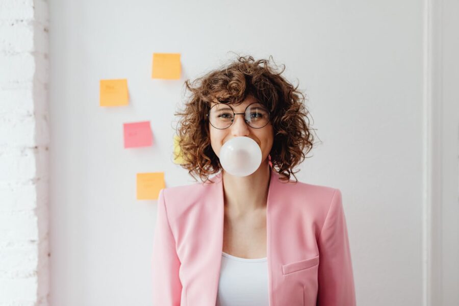 Woman wearing glasses and a pink blazer blowing a large bubble with sugar-free gum to improve breath.